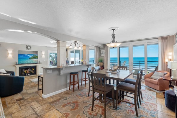 Open-plan living and dining area with ocean view, fireplace, kitchen island, and floor-to-ceiling windows overlooking the beach