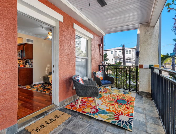 Covered porch with coral textured walls, colorful geometric rug, wicker chairs, and view of palm trees and neighboring buildings
